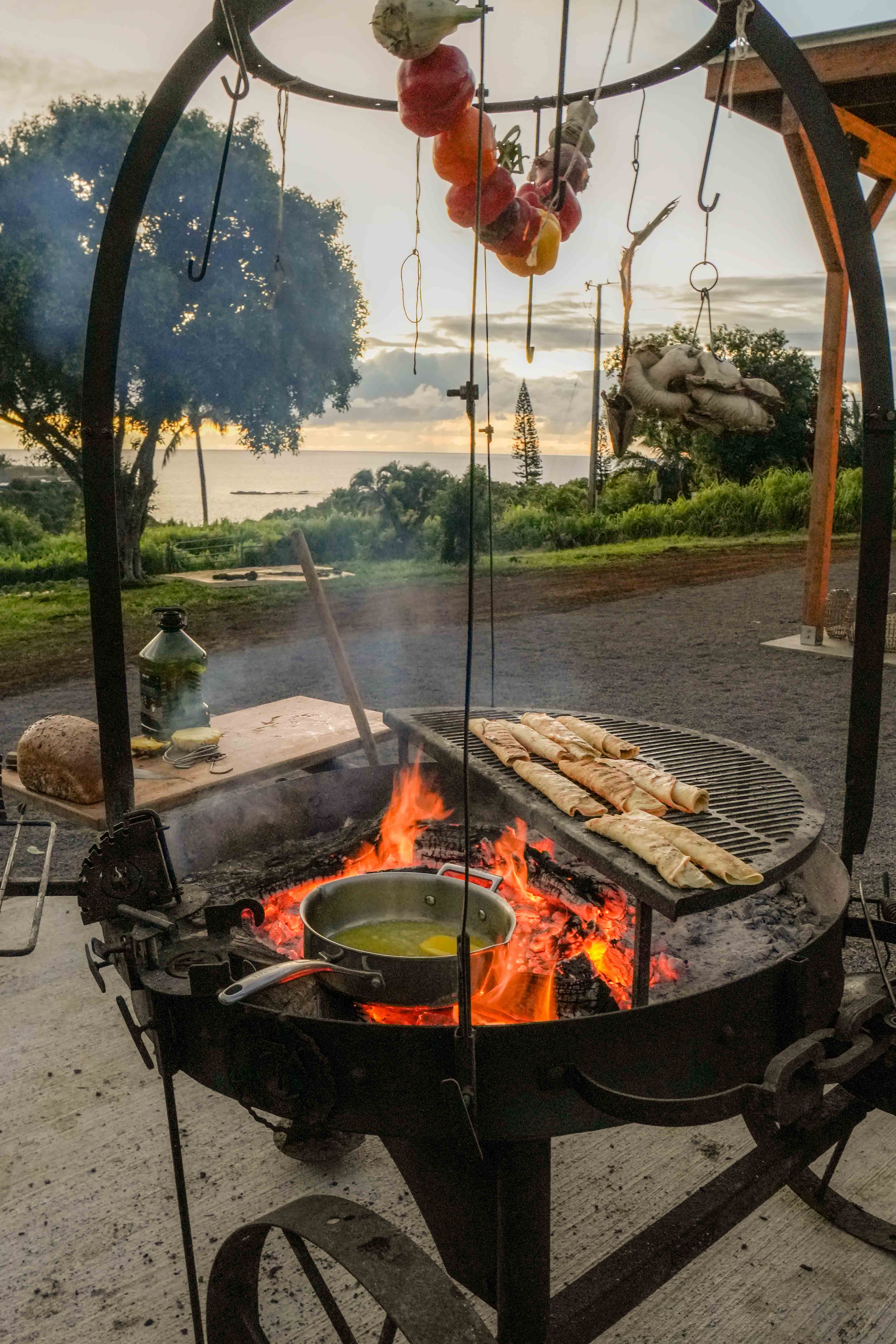 Open-fire grill with hanging peppers and flatbread at sunset over the ocean